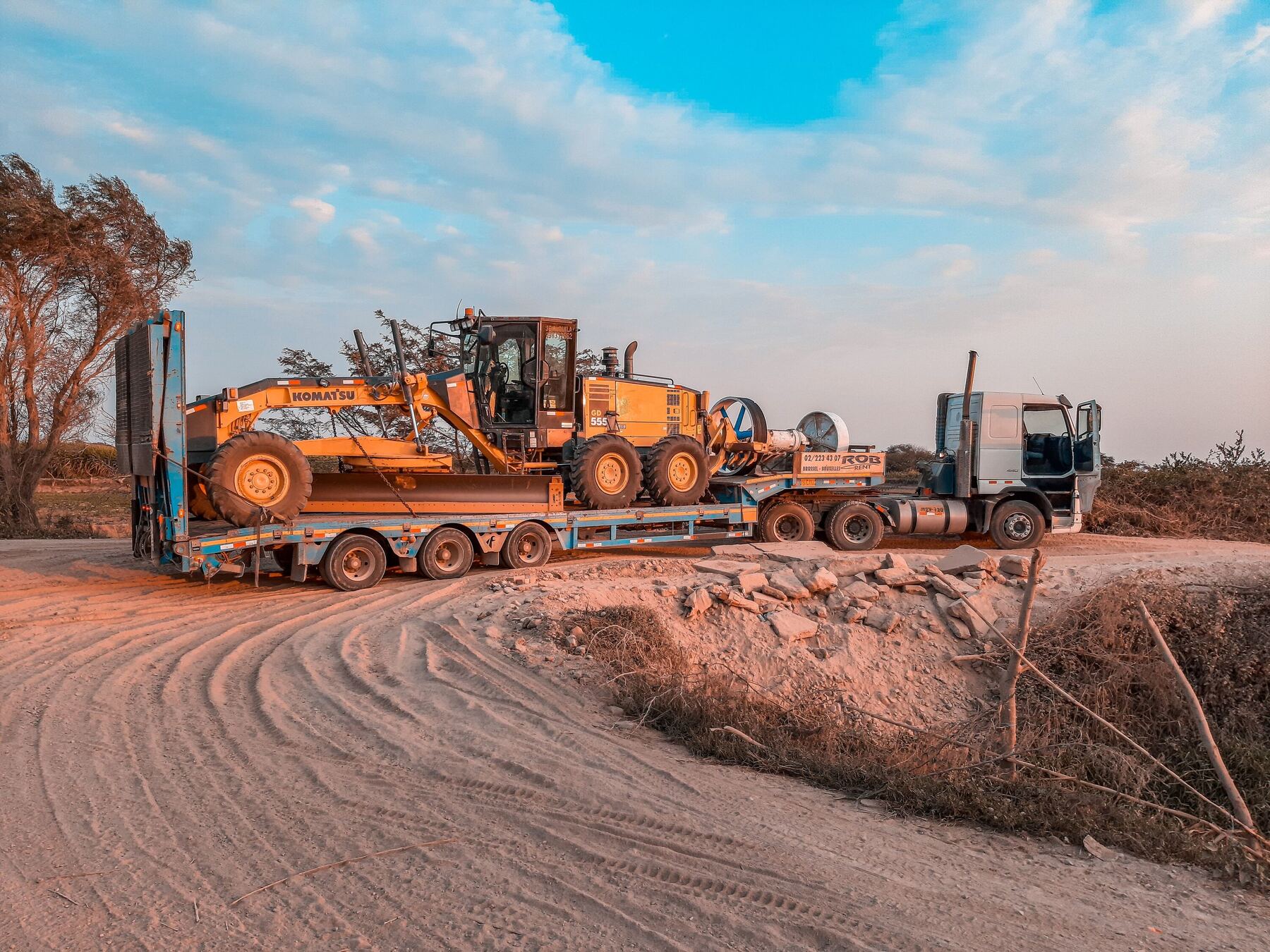 A low-loader trailer carrying a motor grader along a sandy track at golden hour